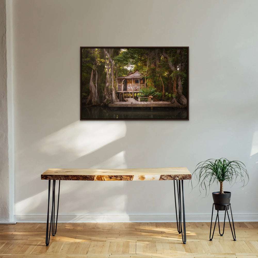 A framed canvas print of "Calypso's House II" by Yuri A Jones on a white wall, hung above a table with a wooden slab top.