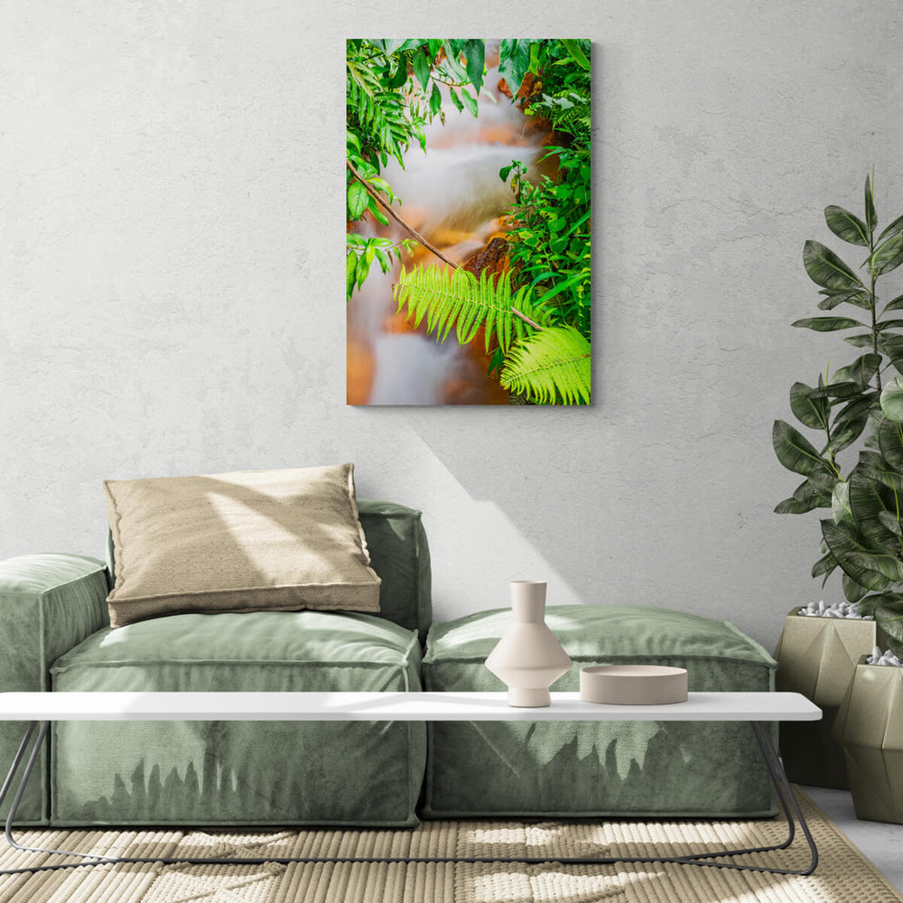 A canvas print of "An Unlikely River II" by Yuri A Jones, hung on a concrete wall, above a green couch with a brown pillow.